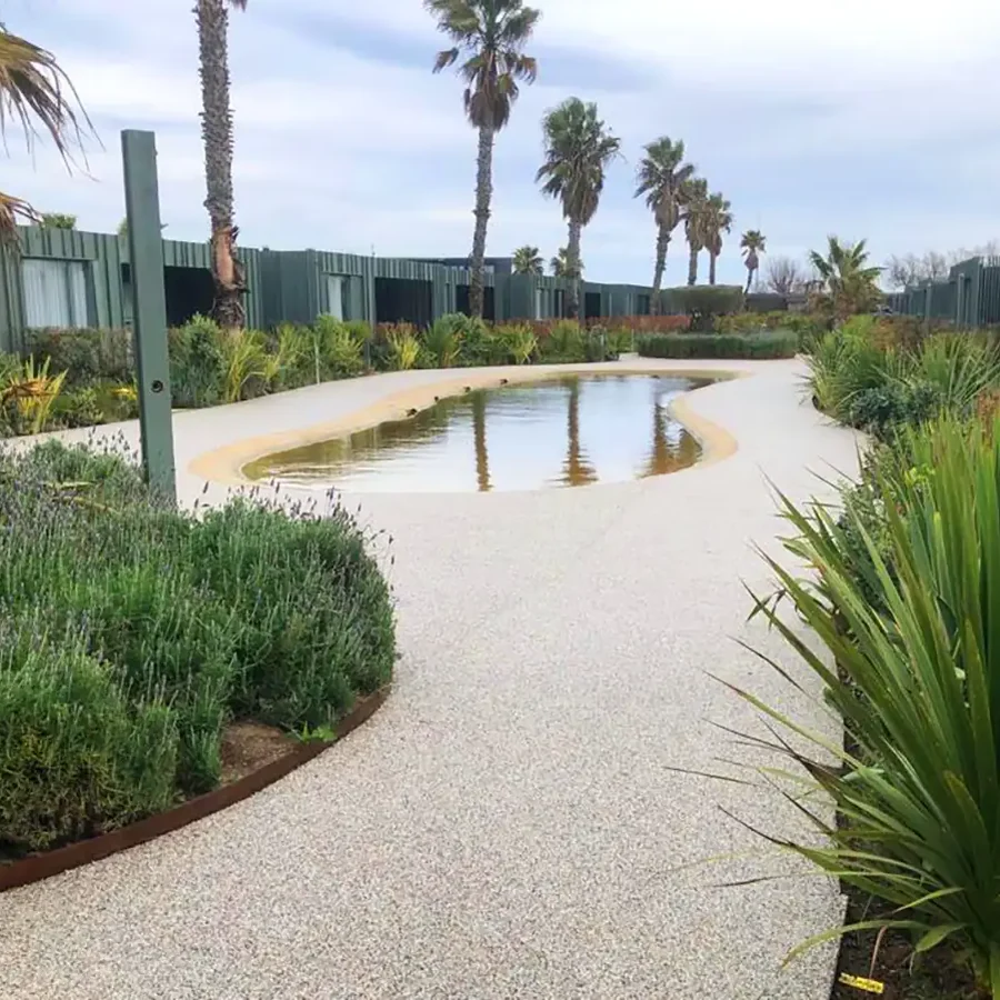Resin-bound crushed-marble path around a small lagoon at Camping La Ballena Alegre, Girona, with palms and dark bungalows