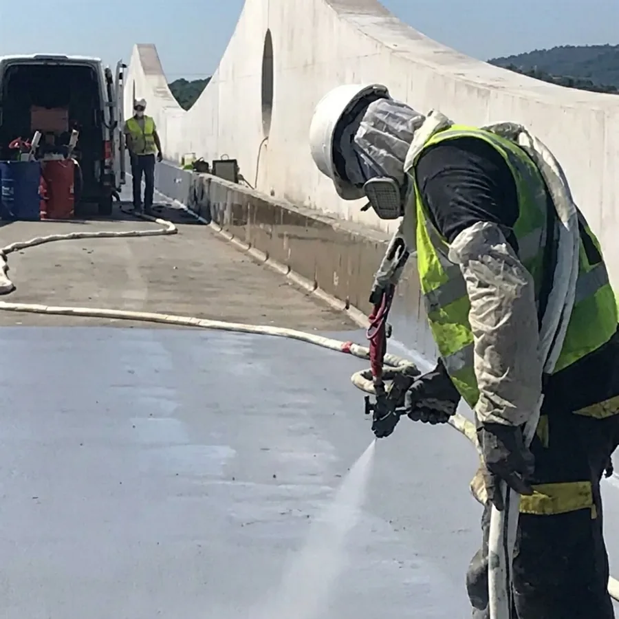 Applicator in a respirator and hi-vis vest hot-spraying a grey membrane on a concrete deck, drums at a van