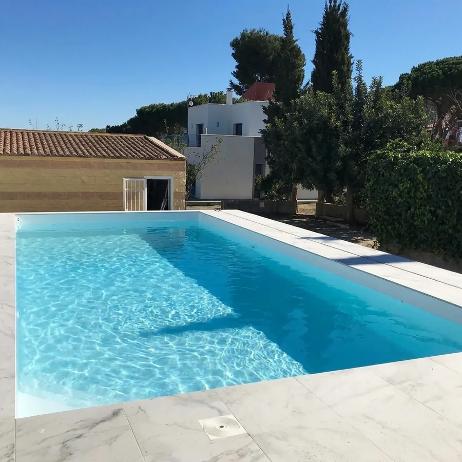 Rectangular rooftop pool filled with blue water, white marble-effect deck tiles, cypress and a terracotta-roof house