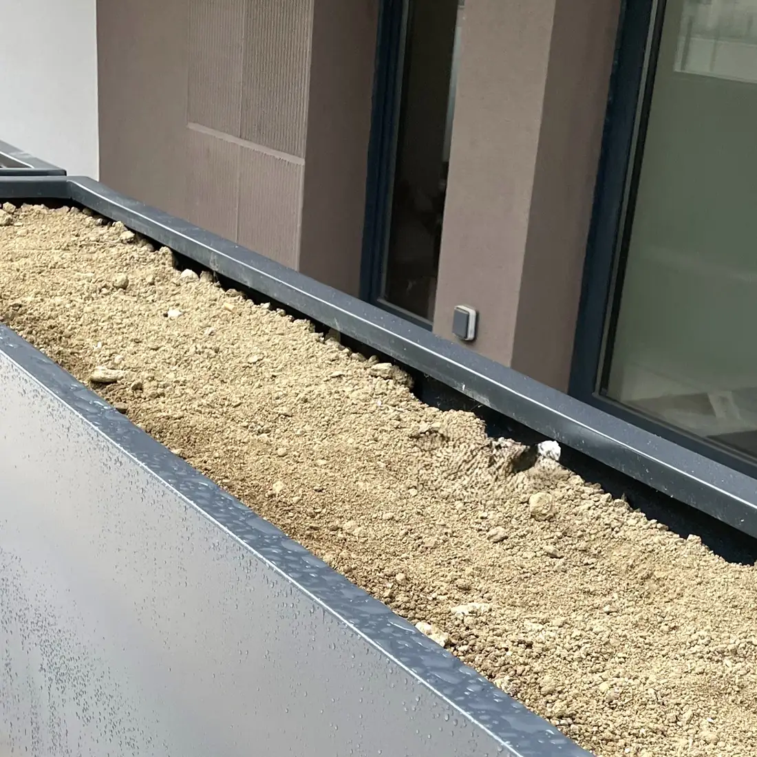 Jardineras metálicas oscuras con grava, junto a una fachada acristalada Jardineras metálicas oscuras con grava, junto a una fachada acristalada